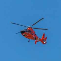 A vibrant red helicopter soaring in a clear blue sky, low-angle view.